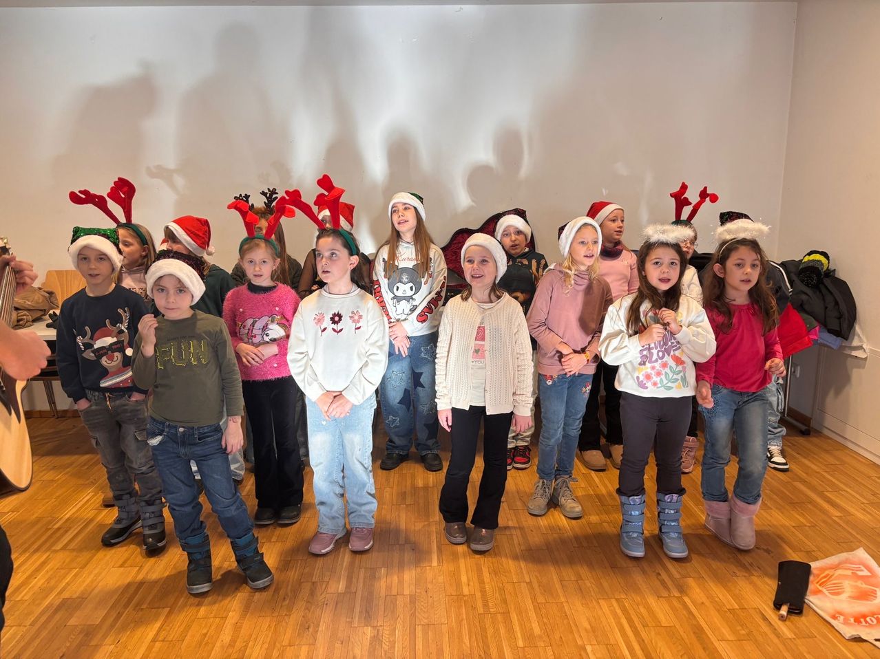 Eine Gruppe von Kindern in Winterkleidung und Weihnachtsmützen steht in einem Raum. Einige tragen Rentier-Kopfbänder und scheinen zu singen.
