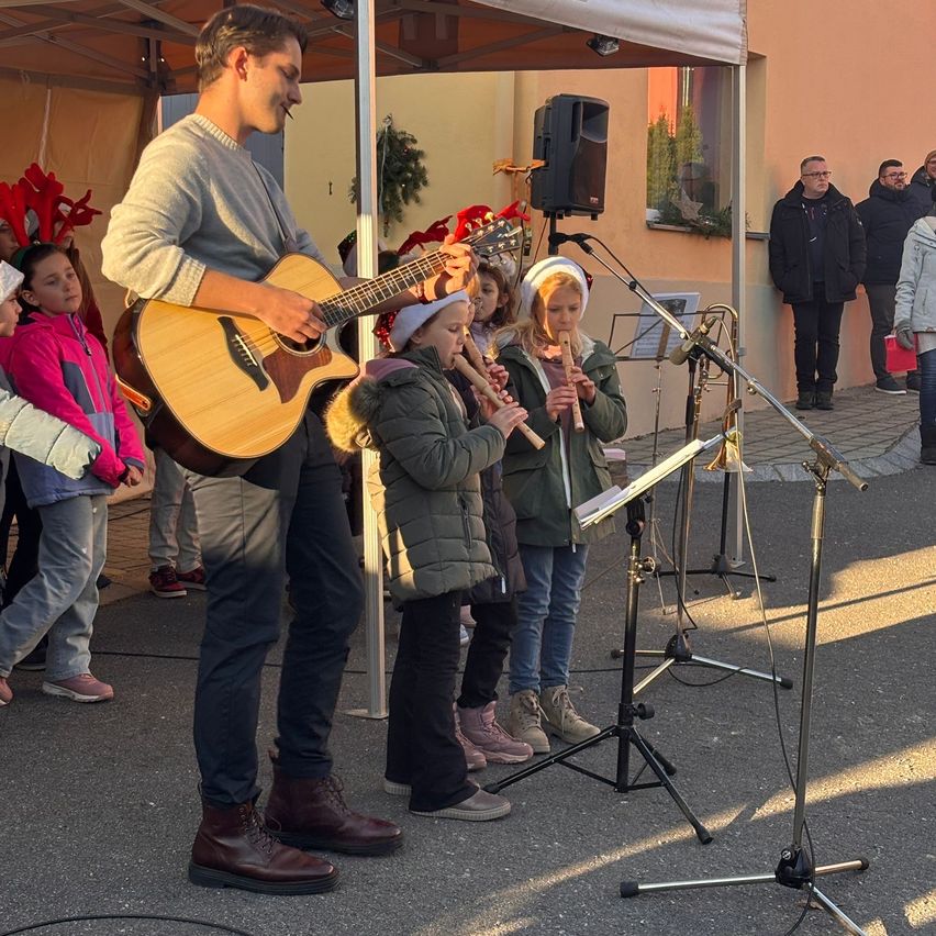 Ein Mann spielt Gitarre unter einem Zelt, während mehrere Kinder Flöten spielen, mit einer Menge, die zusieht. Zwei Männer stehen an der Seite.