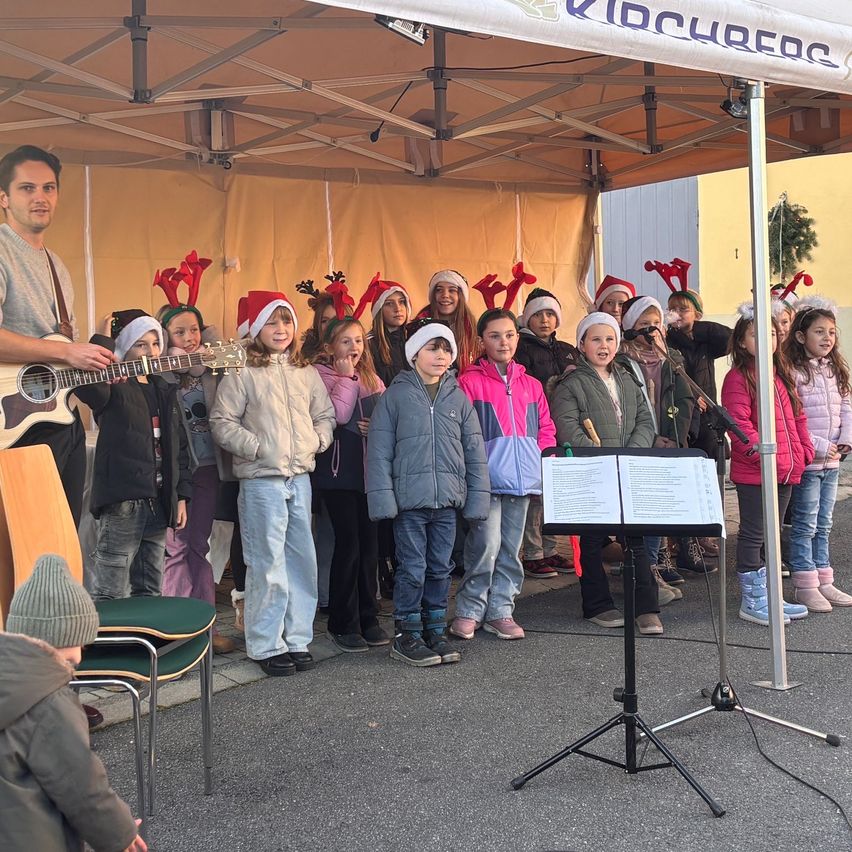 Eine Gruppe von Kindern singt vor einem Zelt mit einem Mann, der Gitarre spielt, alle tragen Weihnachtsmützen.