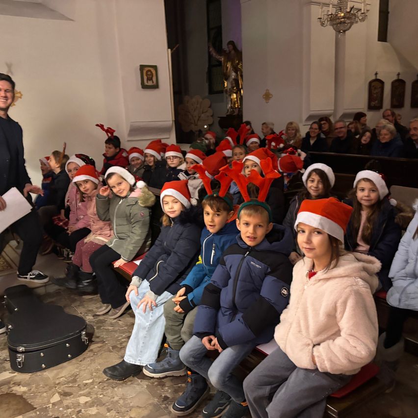 Eine Gruppe von Kindern in Weihnachtsmützen und Jacken sitzen auf Bänken in einer Kirche, vor ihnen steht ein erwachsener Mann, der ein Stück Papier in der Hand hält.