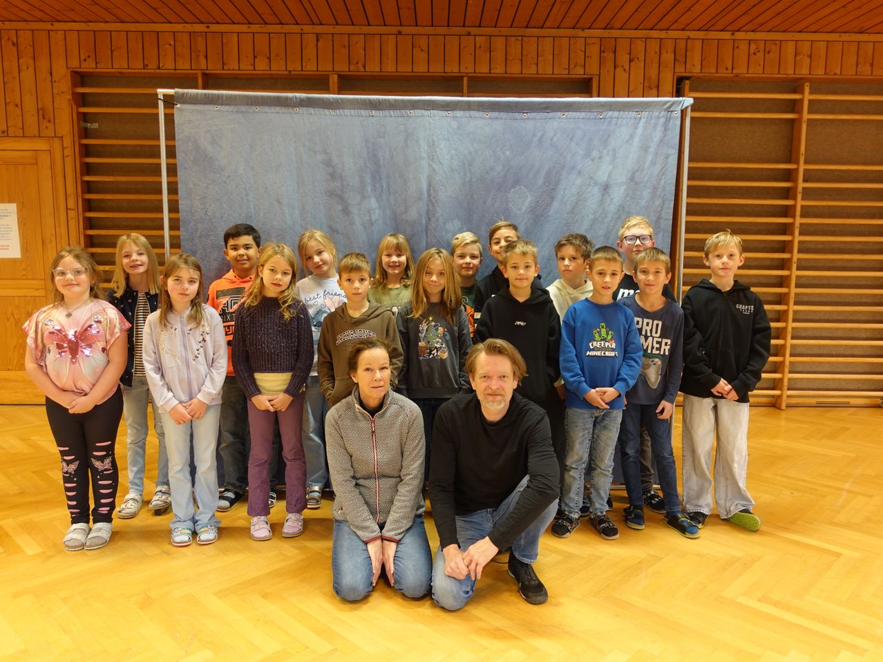 Eine Gruppe von Kindern und zwei Erwachsenen steht vor einem blauen Hintergrund mit einem Banner. Die Kinder tragen Freizeitkleidung, während die Erwachsenen auf dem Boden knien. Das Bild erfasst einen Moment der Einheit und Zusammenarbeit innerhalb der Gruppe.