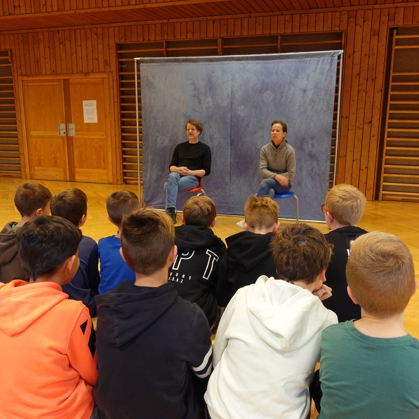 Zwei Personen sitzen auf Hockern vor einem blauen Hintergrund und sprechen zu einer Gruppe junger Jungen, die im Kreis sitzen. Der Raum ist mit Holzwänden gestaltet.