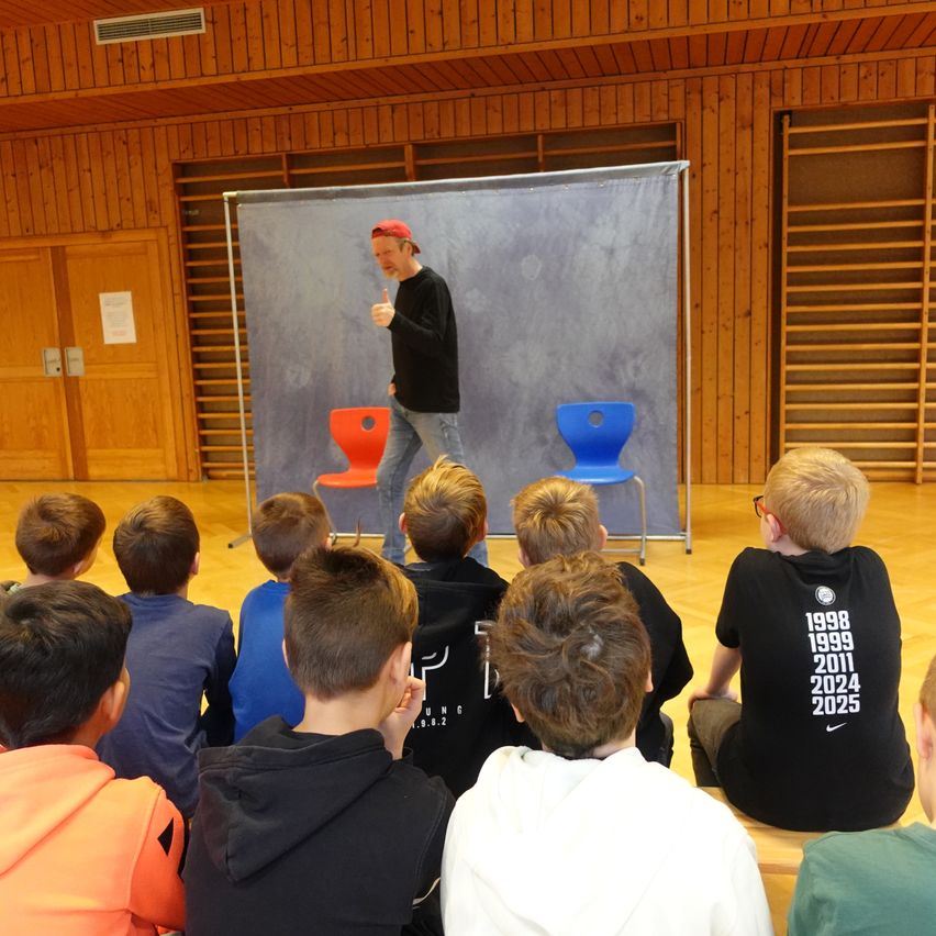 Ein Mann steht auf der Bühne und spricht mit Handgesten zu einer Gruppe von Kindern, die im Halbkreis sitzen. Der Hintergrund ist eine große graue Leinwand mit zwei Stühlen, einem roten und einem blauen.