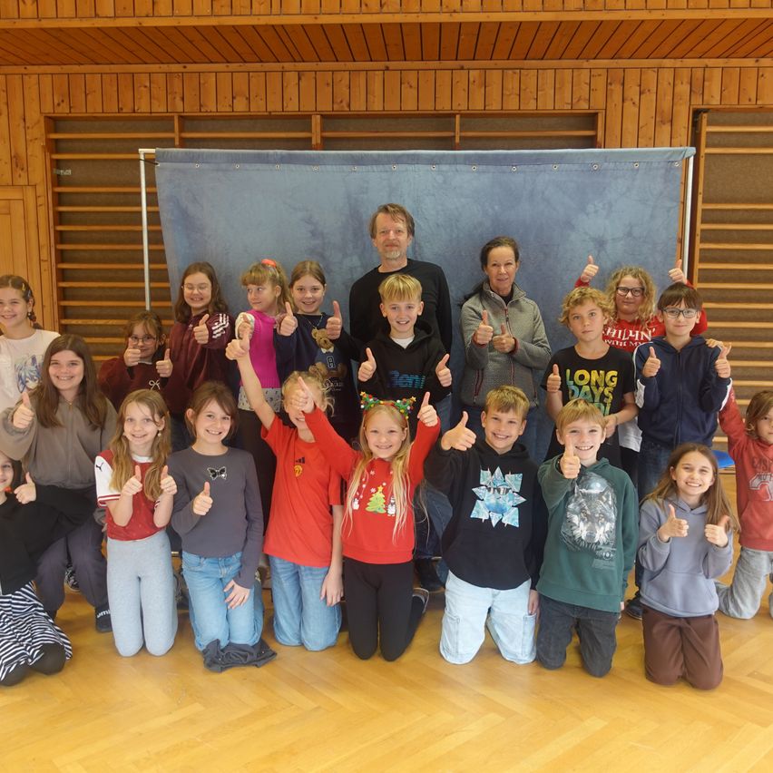 Eine Gruppe von Kindern und zwei Erwachsenen posiert für ein Foto mit erhobenen Daumen in einem Raum mit Holzwänden und -boden.