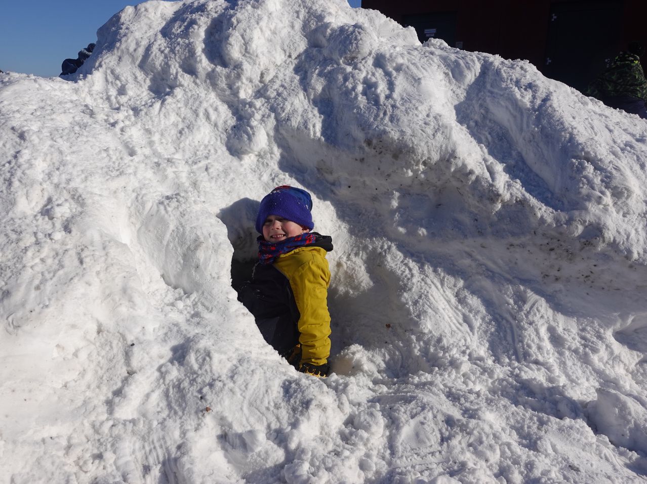 Ein junger Junge in einem gelben Mantel und einer blauen Mütze versteckt sich in einem Schneehaufen.