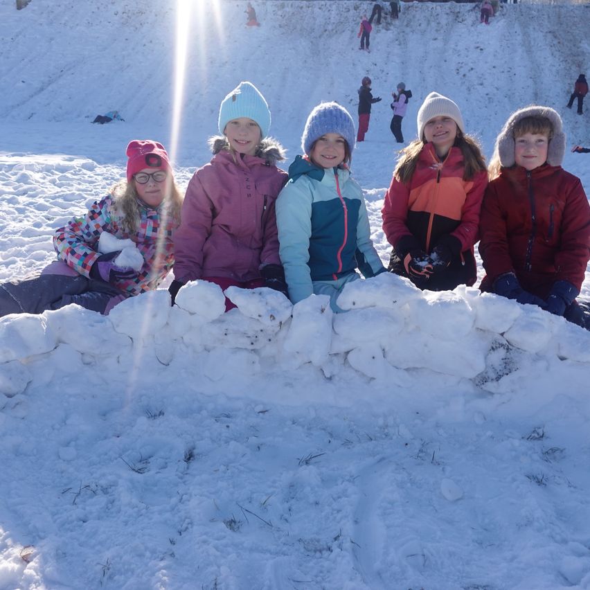 Fünf Kinder posieren für ein Foto auf einem verschneiten Hang, jedes mit Winterjacke und Mütze. Der Schnee ist um sie herum aufgehäuft, mit weiteren Kindern im Hintergrund.