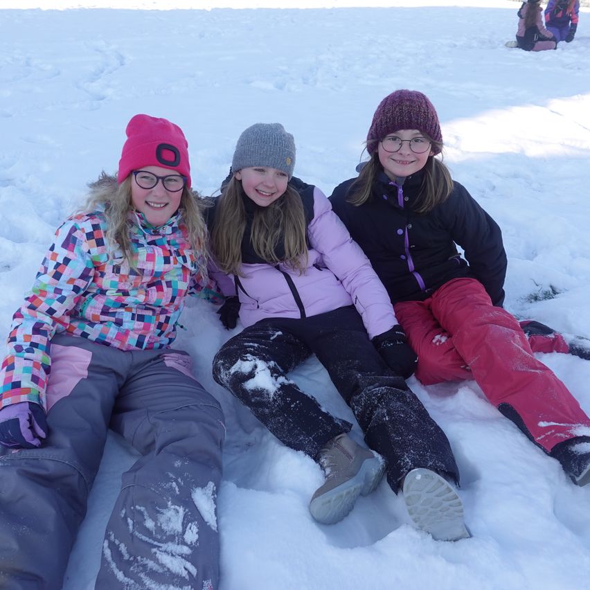 Drei junge Mädchen, warm gekleidet, sitzen im Schnee und lächeln. Sie tragen Mützen und Handschuhe. Schnee liegt auf ihrer Kleidung.