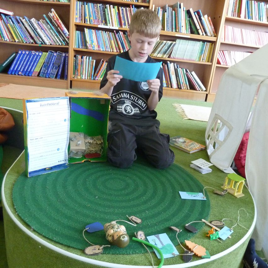 Ein junger Junge sitzt auf einem grünen Teppich und liest ein blaues Papier, mit Büchern und Spielzeug um ihn herum. Er steht vor einem Bücherregal aus Holz, gefüllt mit Büchern.