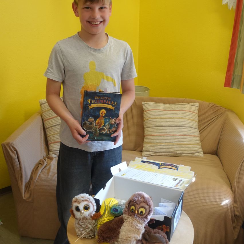 Ein Junge hält ein Buch in den Händen und lächelt. Hinter ihm steht eine Couch mit Kissen. Es gibt einen Karton auf einem Tisch mit Kuscheltieren.