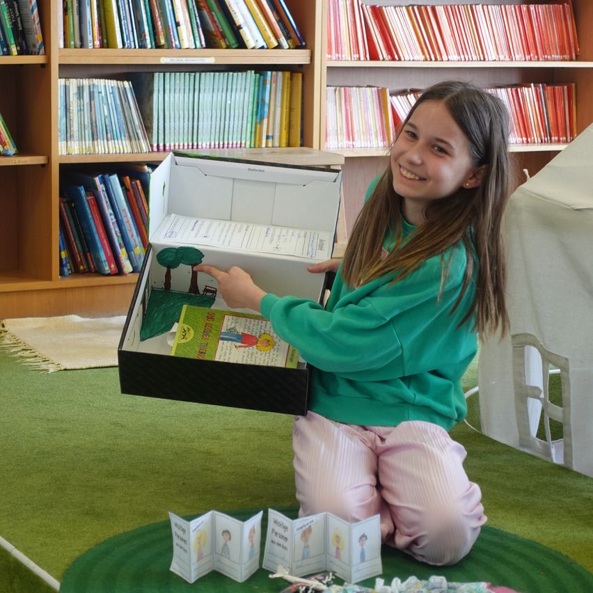 Ein Mädchen sitzt auf dem Boden in einer Bibliothek und hält eine mit Büchern gefüllte Schachtel in der Hand und lächelt.