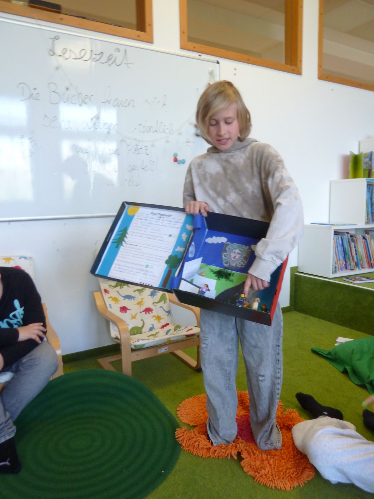 Ein junger Junge steht in einem Klassenzimmer und hält eine offene Box mit einem Lego-Modell. Dahinter steht eine Tafel mit etwas Schrift.