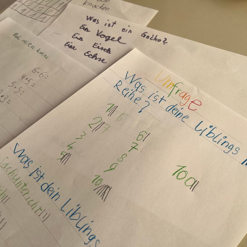 A paper sheet with handwritten mathematical questions is on top of another sheet. The questions are in German and involve arithmetic operations. The paper has numbers and letters written on it.