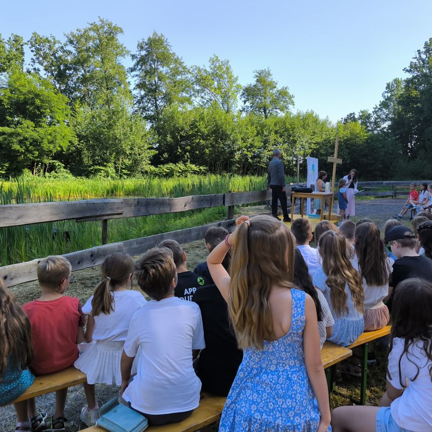 A group of children sit on benches under a wooden fence, watching a man and woman at a table with a cross in front of them. The setting is outdoors with trees and grass in the background.