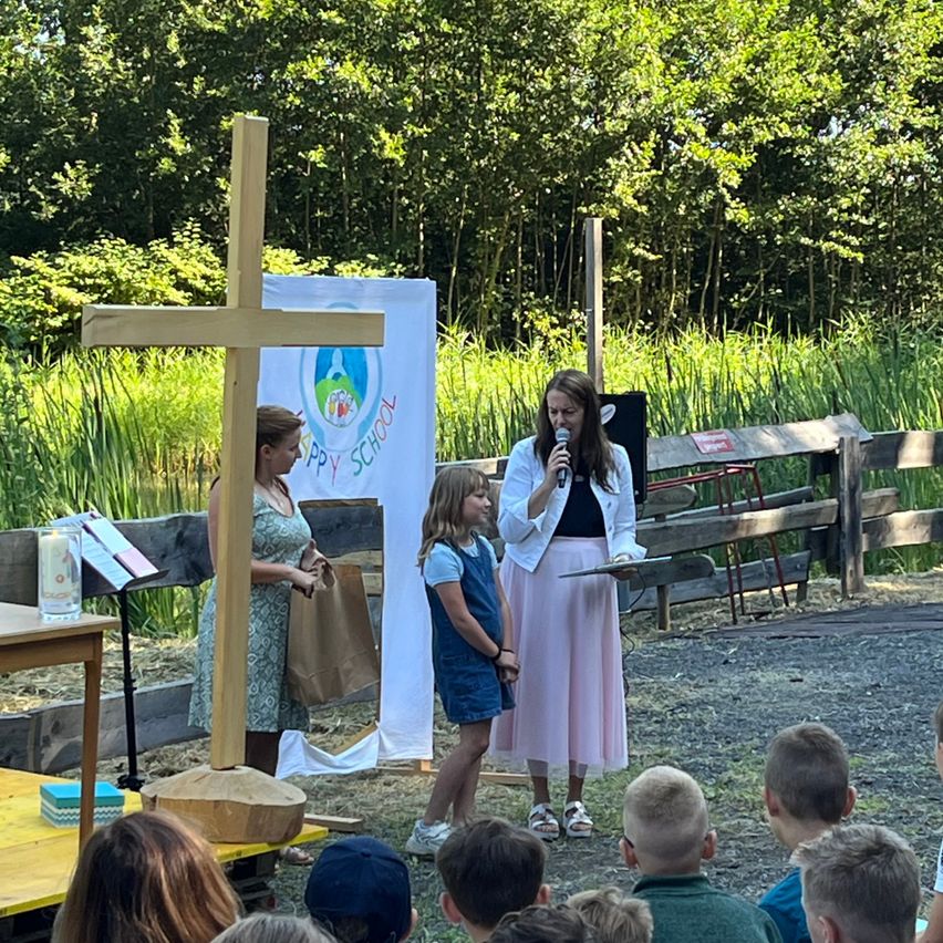 Two women and a girl stand in front of a cross and a banner, speaking to a group of children.