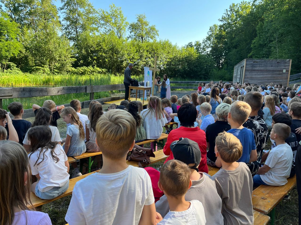A group of children and adults sit on benches in an outdoor setting, watching a man and woman on stage with a wooden cross and table.