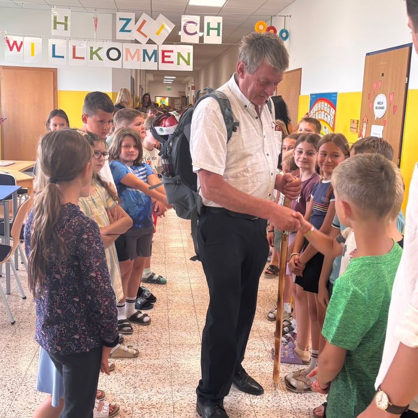 A group of children and an older man with a cane are standing in a hallway with a banner that says 'Willkommen'. The children are looking at the man, and some are holding hands.