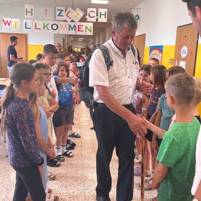 A man with a backpack is shaking hands with a young boy in a hallway with children. The hallway has a banner with the words 'Willkommen' and a colorful wall.