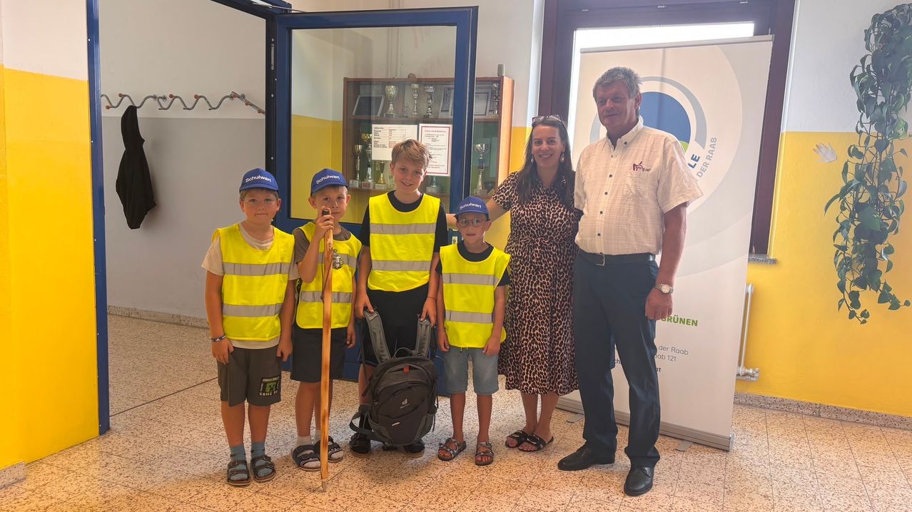 Five children and two adults are standing together, smiling for a photo. The children are wearing yellow safety vests and hats. The man is wearing a belt and a watch. The woman is wearing a leopard-print dress and sandals. They are standing in front of a glass door with a trophy display inside.