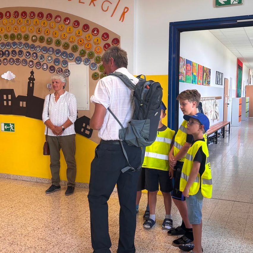 A group of children in yellow vests stand in a hallway with a man and a woman. The hallway has a yellow wall with a mosaic design and an open door.