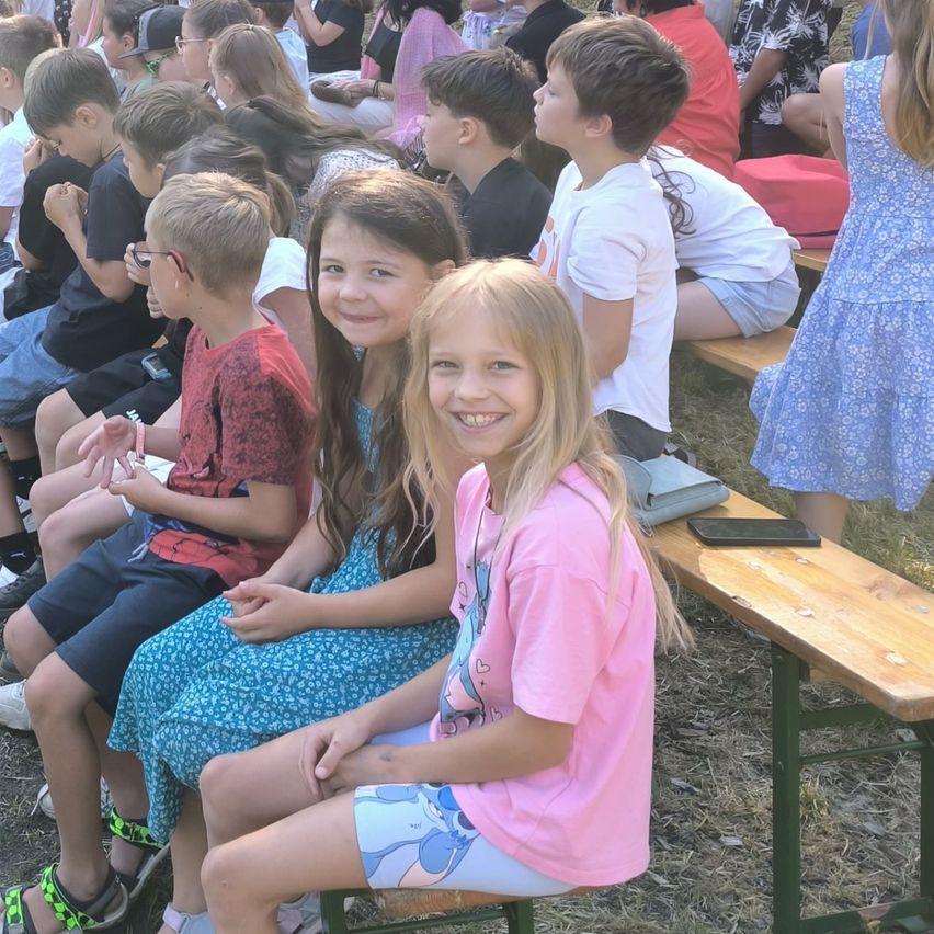 A group of children sit on wooden benches, some smiling and looking at the camera, while others are focused on something in front of them.