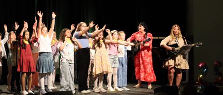 Eine Gruppe von Kindern und zwei Erwachsenen steht auf der Bühne und hebt die Hände. Die Kinder tragen verschiedene Kleidungsstücke, während die Erwachsenen Gitarren halten. Scheinwerfer sind an der Decke sichtbar.