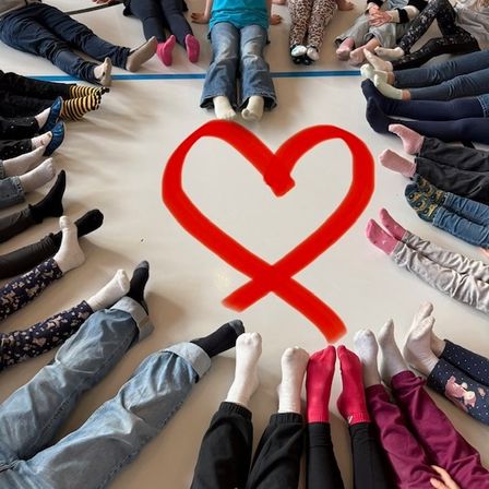 Eine Gruppe von Kindern sitzt im Kreis auf dem Boden und bildet mit ihren Füßen eine Herzform. Sie tragen Socken und einige sind barfuß. In der Mitte des Herzens befindet sich ein rotes Band.