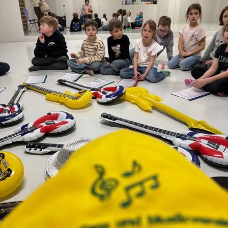 Kinder sitzen im Kreis um aufblasbare Gitarren. Einige lesen Noten. Ein gelber Hut mit Musiknoten ist im Vordergrund.