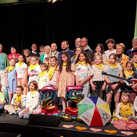 Eine Gruppe von Menschen, einschließlich Kindern, steht auf einer Bühne mit Luftballons und einem Regenschirm. Sie tragen alle gelbe Schals. Einige halten Gitarren. Die Bühne hat einen schwarzen Hintergrund.