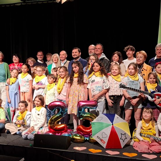 Eine Gruppe von Menschen, einschließlich Kindern, steht auf einer Bühne mit Luftballons und einem Regenschirm. Sie tragen alle gelbe Schals. Einige halten Gitarren. Die Bühne hat einen schwarzen Hintergrund.