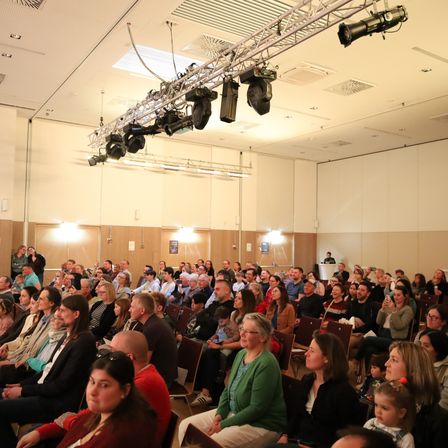 Eine große Menschenmenge sitzt in Stühlen in einem Konferenzsaal mit Bühnenbeleuchtungsgeräten, die von der Decke hängen.