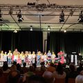 Eine Gruppe von Kindern steht mit Luftballons vor sich auf der Bühne und führt für ein großes Publikum auf. Eine Person spielt Klavier und eine andere steht neben einem Mikrofon. Scheinwerfer hängen an der Decke und schwarze Vorhänge sind hinter der Bühne.