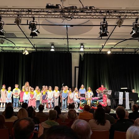 Eine Gruppe von Kindern steht mit Luftballons vor sich auf der Bühne und führt für ein großes Publikum auf. Eine Person spielt Klavier und eine andere steht neben einem Mikrofon. Scheinwerfer hängen an der Decke und schwarze Vorhänge sind hinter der Bühne.