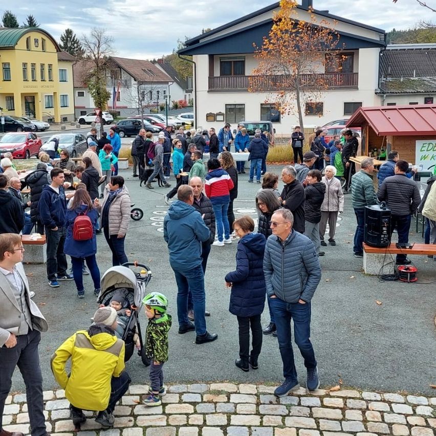 Eine Menschenmenge versammelt sich vor einem Gebäude, einige in Jacken und Turnschuhen. In der Nähe sind Autos geparkt, und es gibt Tische mit Essen. Ein Mann mit Brille und Jacke steht links, und ein Mann in einem Anzug kniet, um mit einem Kind zu interagieren.