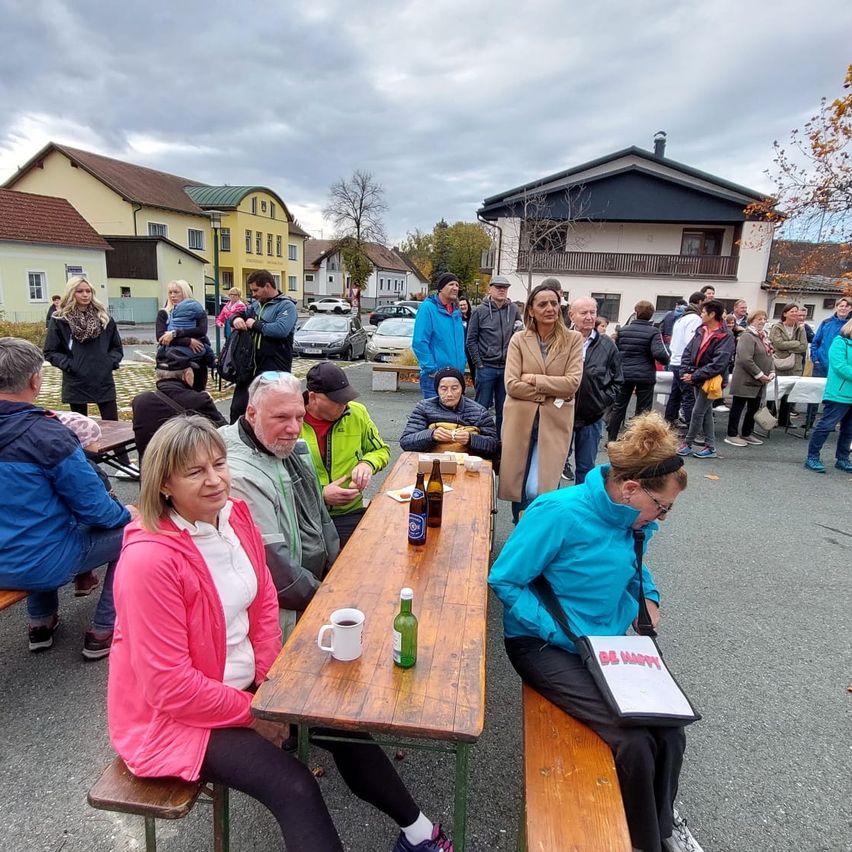 Eine Gruppe von Menschen sitzt an einem Picknicktisch im Freien, mit einem bewölkten Himmel darüber. Der Tisch ist mit Essen und Getränken beladen. In der Nähe stehen, gehen und unterhalten sich mehrere Personen. Im Hintergrund sind Häuser und Autos zu sehen.