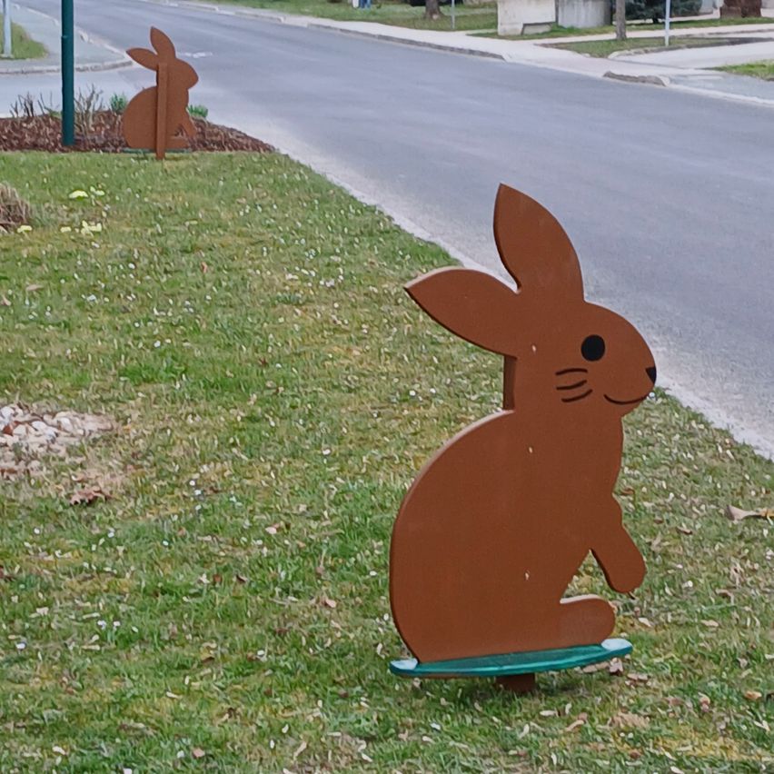 A brown rabbit figurine sits on a green skateboard on the grass by a road. Another rabbit stands in the background. The road is empty.