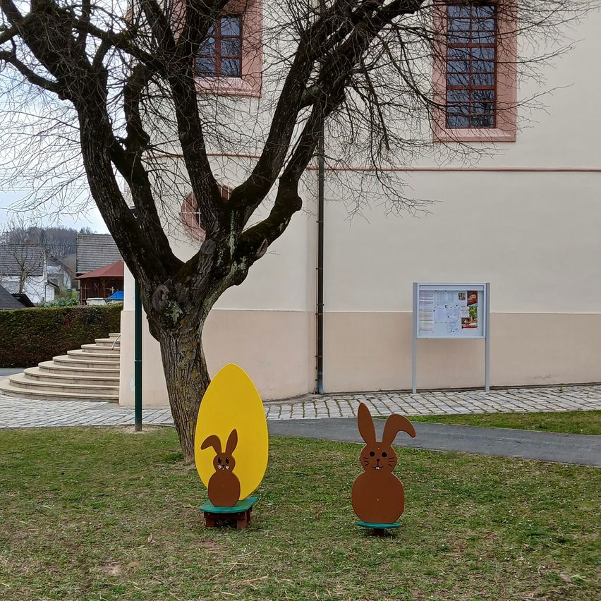 Two wooden Easter decorations stand in front of a building with windows, a tree, and stairs. One is an egg with a rabbit, and the other is a rabbit on a small stand.