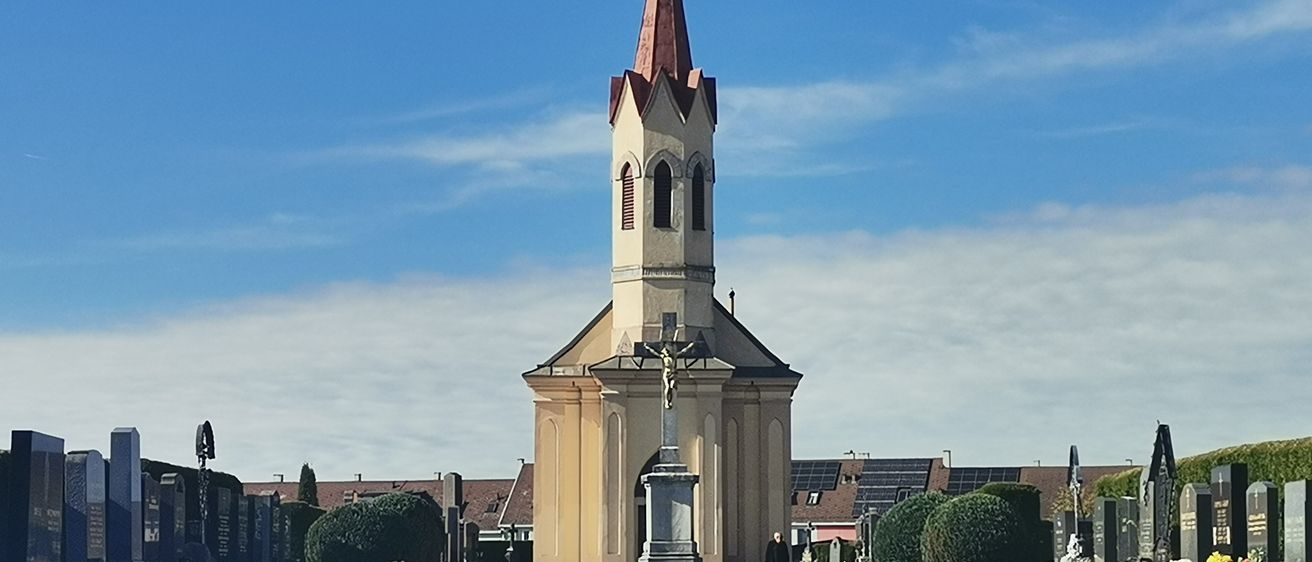 Eine Kirche mit einem Turm und einem Kreuz steht auf einem Friedhof, umgeben von einem Weg, Grabsteinen und Pflanzen. Solarmodule sind auf den Dächern der Gebäude im Hintergrund zu sehen.