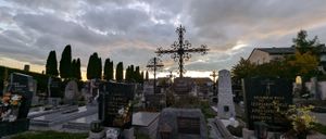 Ein Friedhof mit mehreren Grabsteinen und Kreuzen. Der Himmel ist bewölkt und die Sonne geht unter. Es gibt ein großes Steinkreuz mit einer runden Platte in der Mitte.
