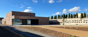 Ein modernes Gebäude mit Glaswänden steht unter einem blauen Himmel mit Wolken. Es hat eine Einfahrt vor dem Gebäude mit Erde und Pflanzen. Hohe Bäume stehen hinter dem Gebäude.