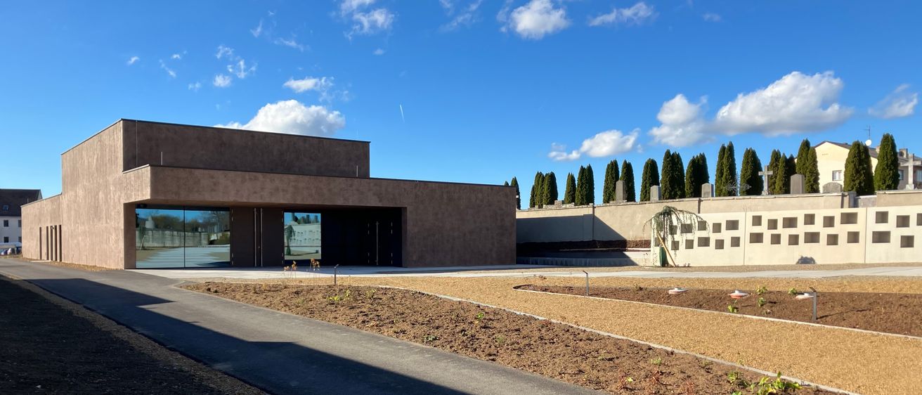 Ein modernes Gebäude mit Glaswänden steht unter einem blauen Himmel mit Wolken. Es hat eine Einfahrt vor dem Gebäude mit Erde und Pflanzen. Hohe Bäume stehen hinter dem Gebäude.