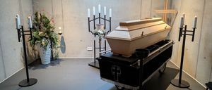 Ein Raum mit einer Betonwand, einem rollenden Wagen mit einem Sarg und einem Kreuz oben, mit Blumen und Kerzen an der Seite.