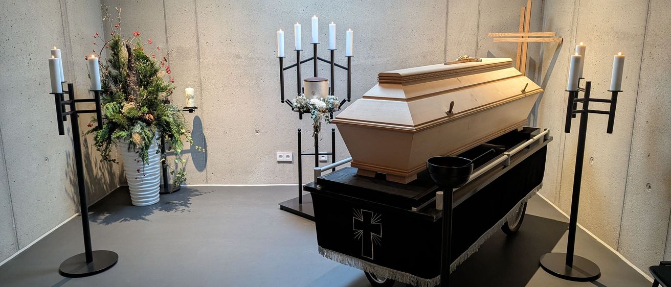 Ein Raum mit einer Betonwand, einem rollenden Wagen mit einem Sarg und einem Kreuz oben, mit Blumen und Kerzen an der Seite.