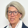 An older woman with short blonde hair wearing glasses and a scarf. She is smiling slightly and wearing a blue jacket.