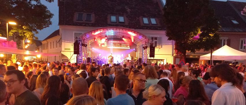 Ein Outdoor-Musikereignis mit einer Menge, die die Show vor einem Gebäude mit roten Lichtern genießt. Eine Bühne mit Musikern, die auftreten.