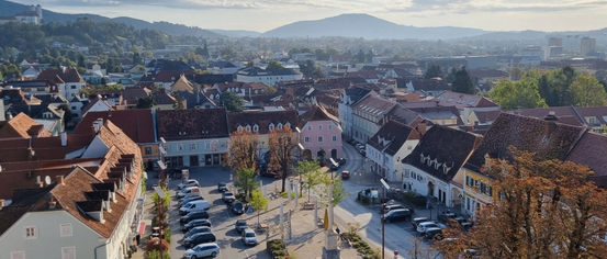 Ein Stadtplatz mit mehreren Gebäuden, geparkten Autos und einem Denkmal in der Mitte. Bäume und Berge sind in der Ferne.