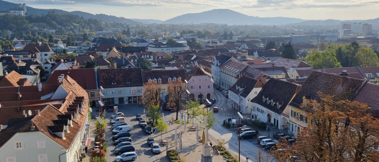 Ein Stadtplatz mit mehreren Gebäuden, geparkten Autos und einem Denkmal in der Mitte. Bäume und Berge sind in der Ferne.