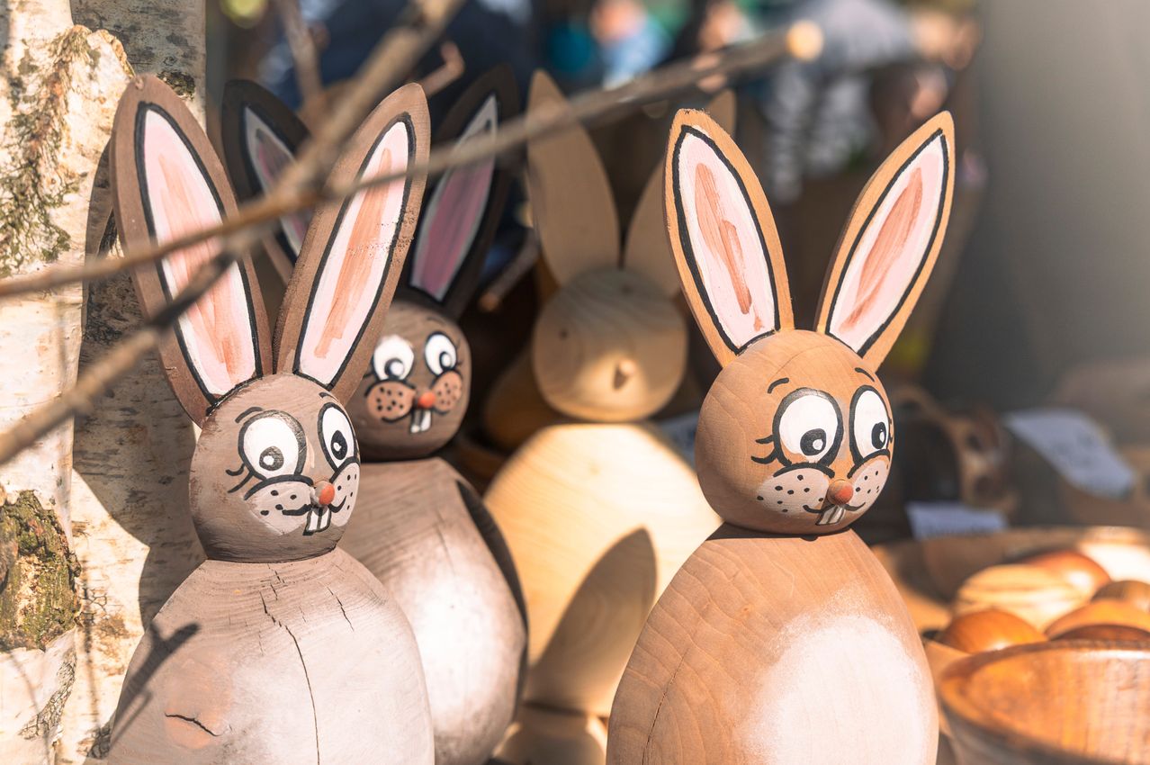 Ein Nahaufnahme von einer Sammlung von Holzhasen. Die Hasen haben gemalte Augen, Ohren und Nasen. Der Hintergrund ist unscharf, mit Menschen und Zweigen im Hintergrund. Die Schatten der Hasen werden auf den Boden geworfen.