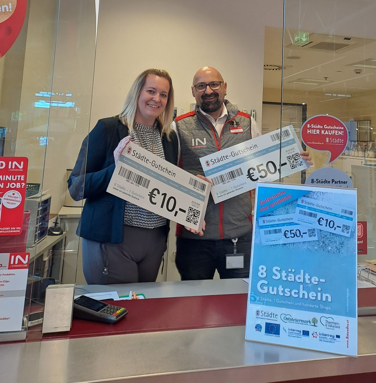 Zwei Personen stehen hinter einem Tresen und halten einen 10-Euro- und einen 50-Euro-Gutschein. Die Frau trägt eine blaue Jacke und der Mann einen roten und grauen Weste. Rechts befindet sich ein Schild mit einem QR-Code.