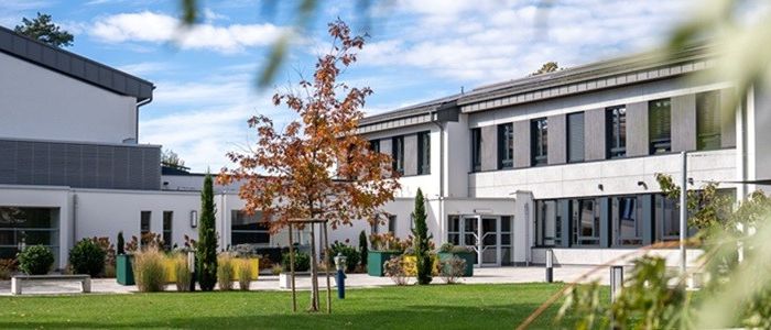 Ein Bild eines modernen Gebäudes mit einem Garten davor. Das Gebäude hat Glasfenster und eine Glastür. Bäume und Pflanzen umgeben das Gebiet.