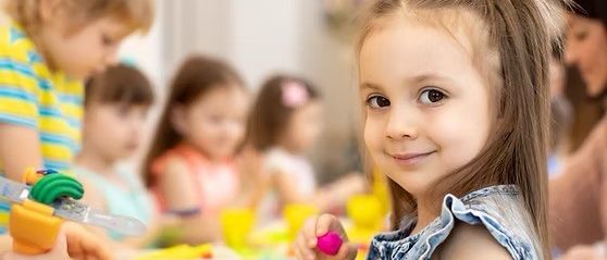 Ein kleines Mädchen lächelt, während es ein rosa Objekt hält, umgeben von anderen Kindern, die an einer Bastelaktivität teilnehmen.